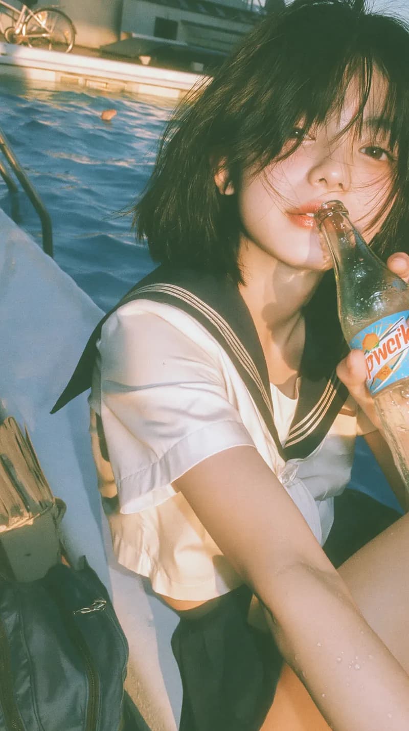 A short-haired figure holding a soda by a sunlit pool, with a grainy film texture